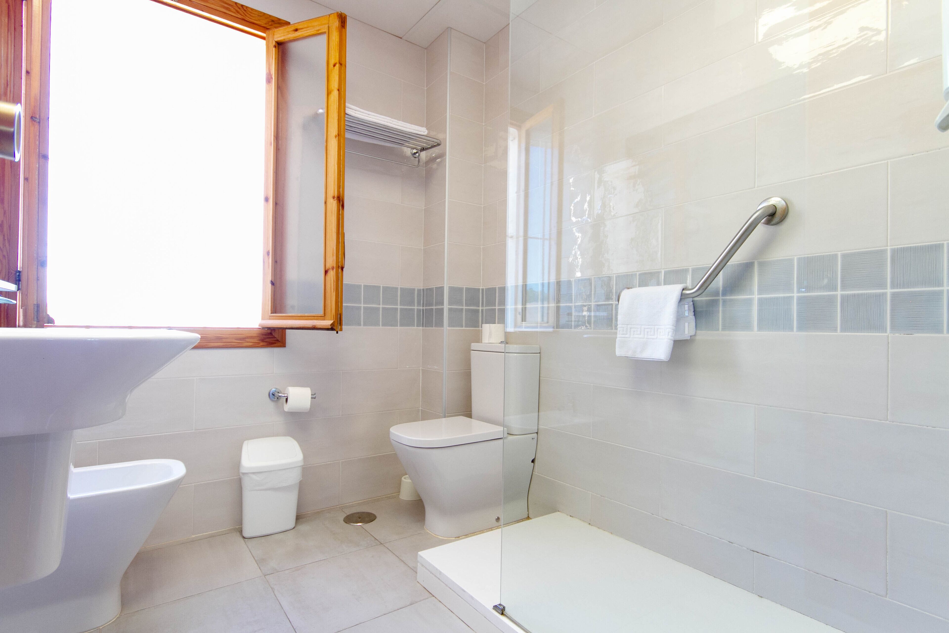 Baño con lavamanos, inodoro y ducha blancos. Hay un perchero en la pared y una ventana sobre el inodoro.