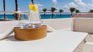 Una mesa con un vaso de agua y una bebida sobre un posavasos de madera, con vistas a una playa con palmeras y el océano.