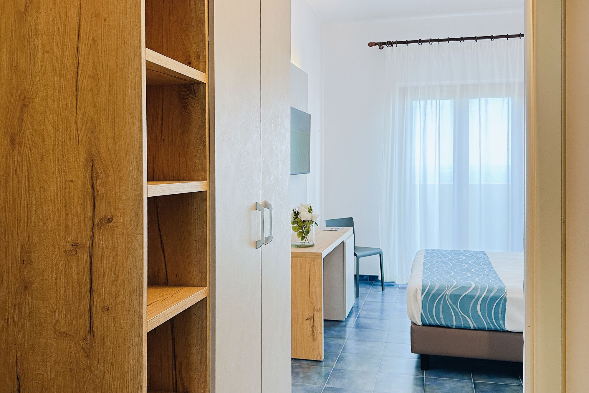 A bedroom with a bed, a desk, and a chair. There is a wooden wardrobe and a window with curtains.