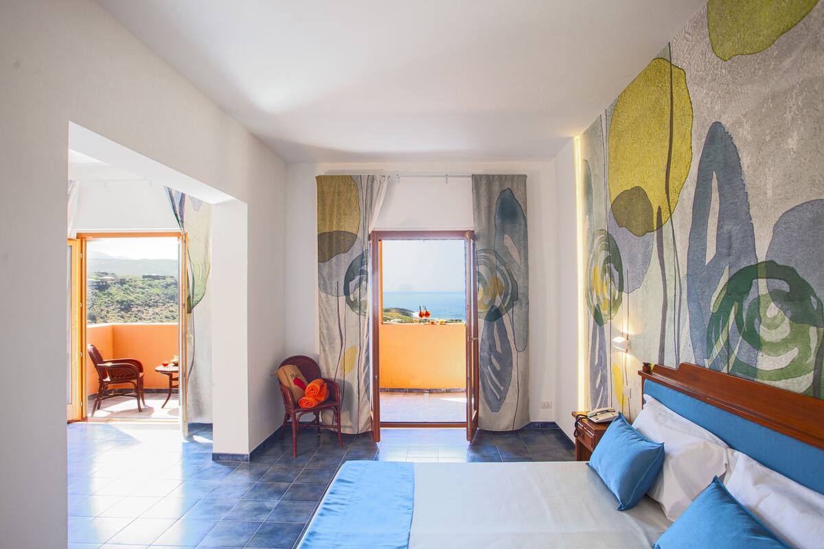 A hotel room with a bed, a chair, a table, and a view of the sea through a balcony.