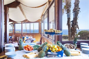 A dining area with a table set for a meal, featuring a variety of fruits and dishes, overlooking the ocean.
