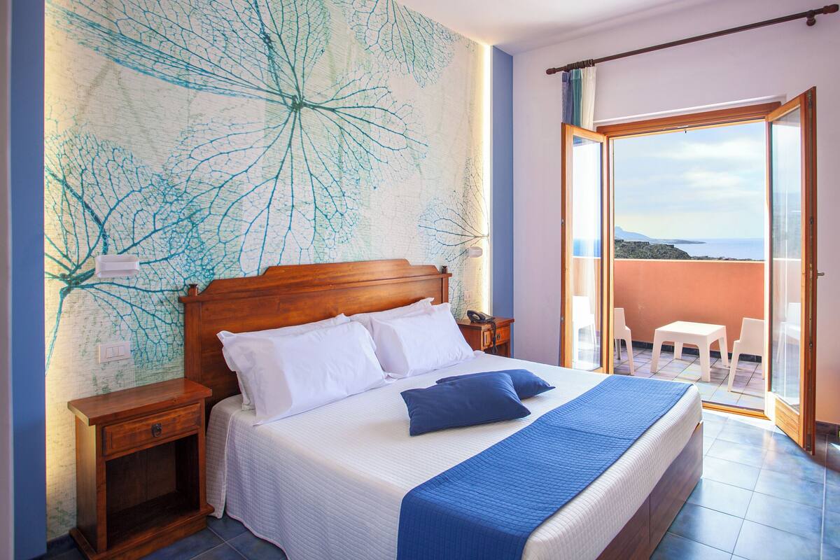 A bedroom with a wooden headboard, white bedding, and a view of the sea through an open balcony door.