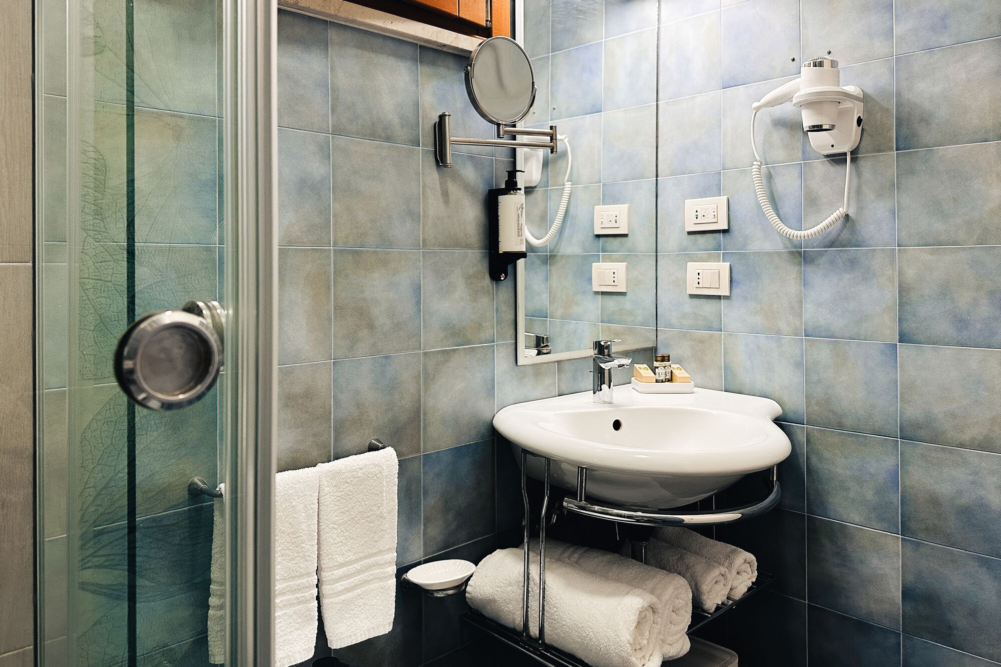 A bathroom with a glass shower enclosure, a white sink with a mirror above it, and a towel rack with white and gray towels.