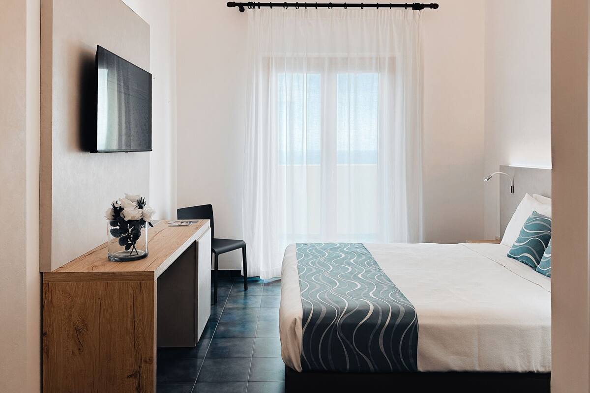 A hotel room with a bed, a desk, a chair, a TV mounted on the wall, and a window with sheer curtains.