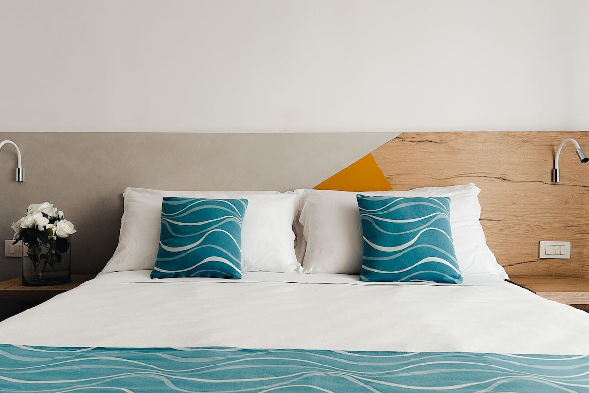 A neatly made bed with white bedding and two teal pillows, a nightstand with a vase of white flowers, and a wooden headboard with a geometric design.
