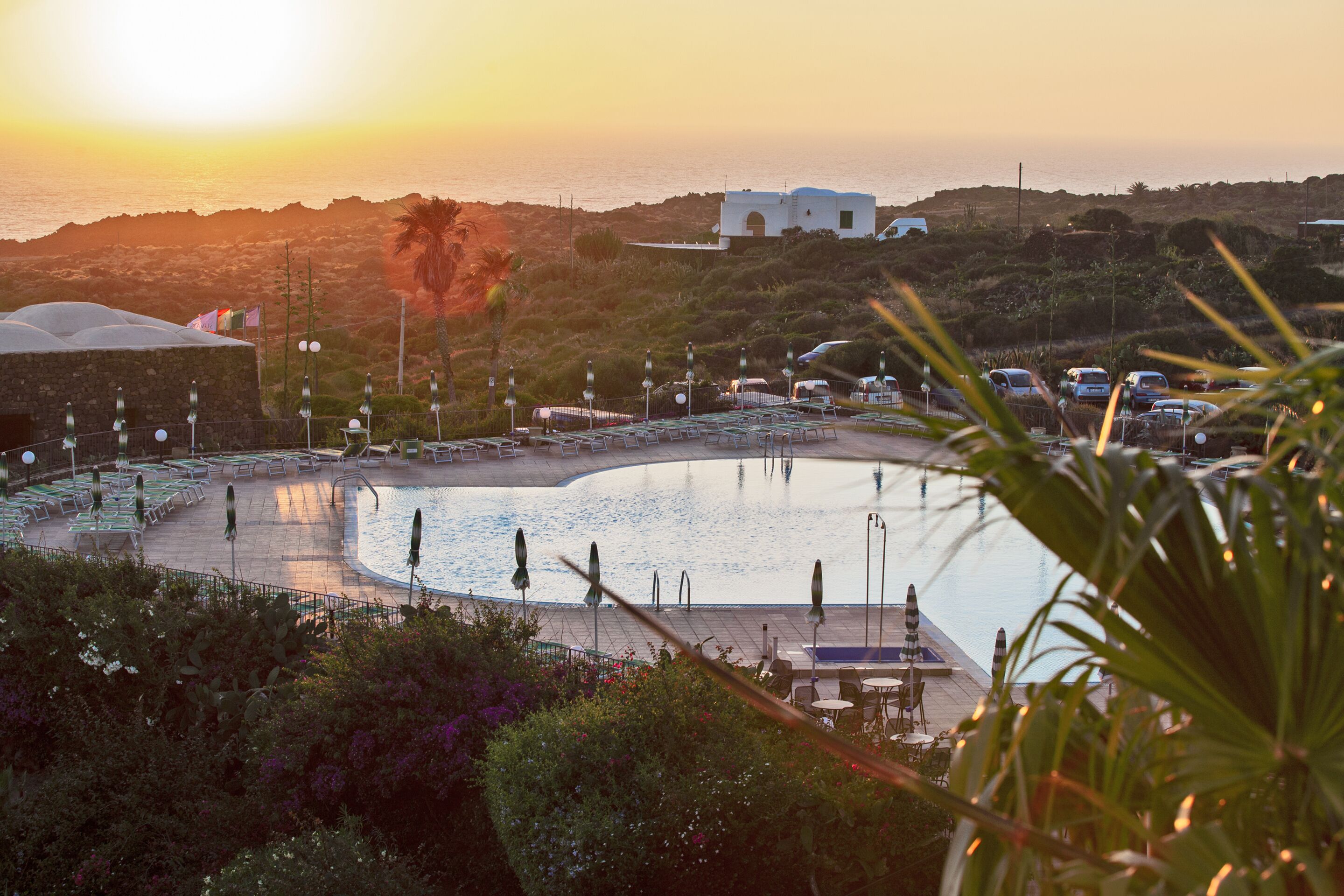 Un área de piscina con sillones de descanso y sombrillas, rodeada de vegetación y con vistas a una masa de agua al atardecer.