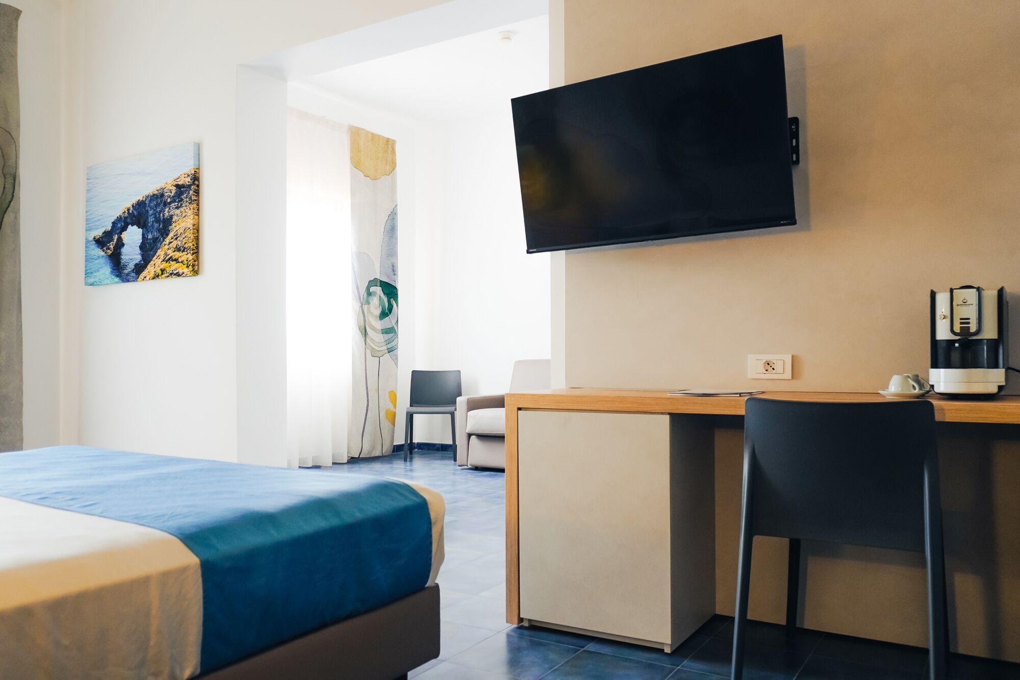 A hotel room with a bed, a desk, a chair, a television, and a painting on the wall.