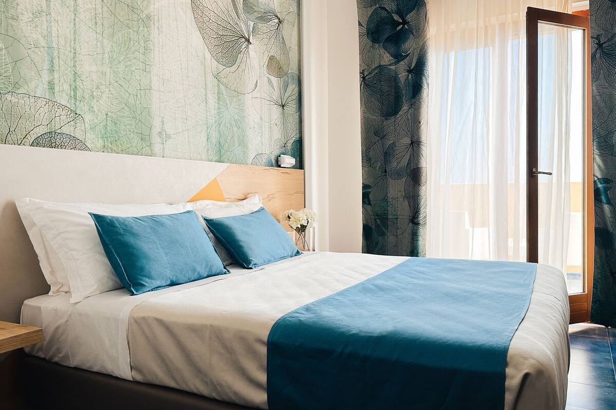 A neatly made bed with blue and white bedding, a wooden headboard, and a view of the outdoors through a glass door.