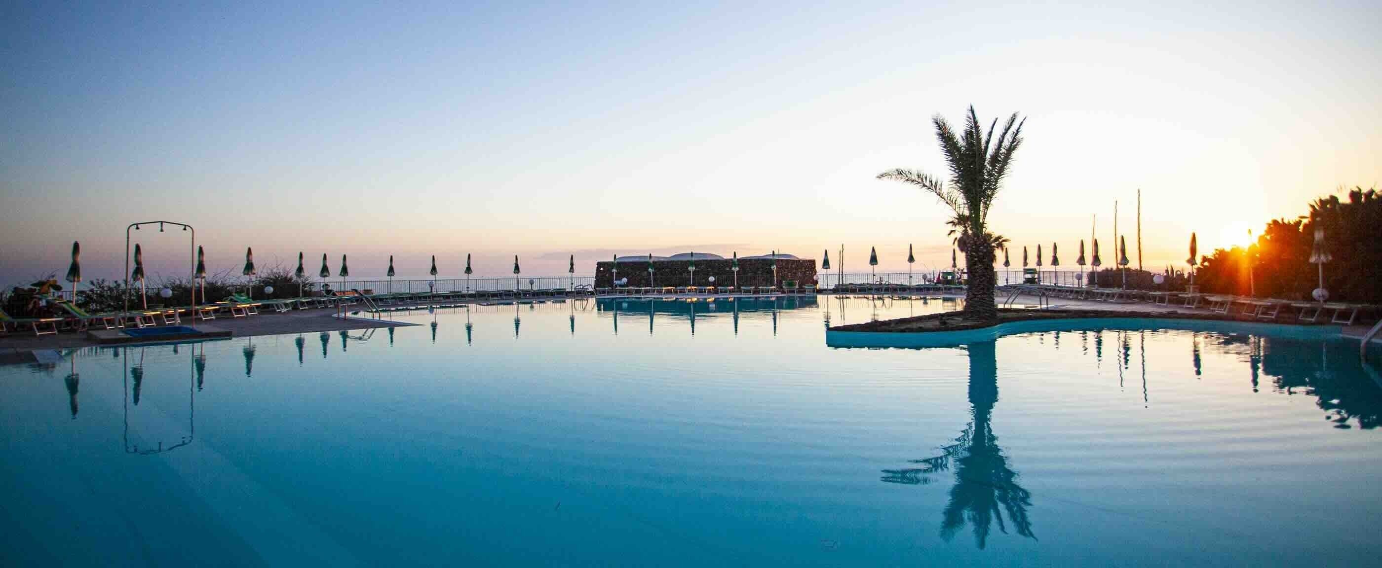 A calm pool area with lounge chairs and umbrellas, surrounded by a fence and palm trees.