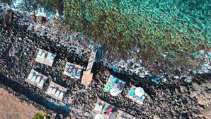 Luchtbeeld van een strandresort met meerdere houten terrassen, helder turquoise water en rotsachtige oevers.