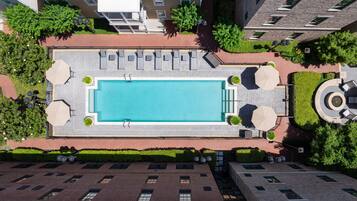 An aerial view of a rooftop swimming pool with lounge chairs and umbrellas, surrounded by greenery and adjacent buildings.