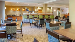 Un comedor de hotel con varias mesas y sillas, una barra con dispensador de bebidas y una pared decorada con obras enmarcadas.