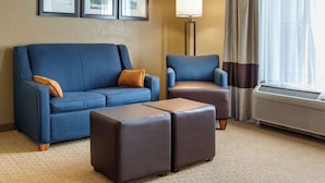 Habitación de hotel con sofá azul, sillón marrón, dos otomanas a juego, una lámpara de pie y un ventanal con cortinas.