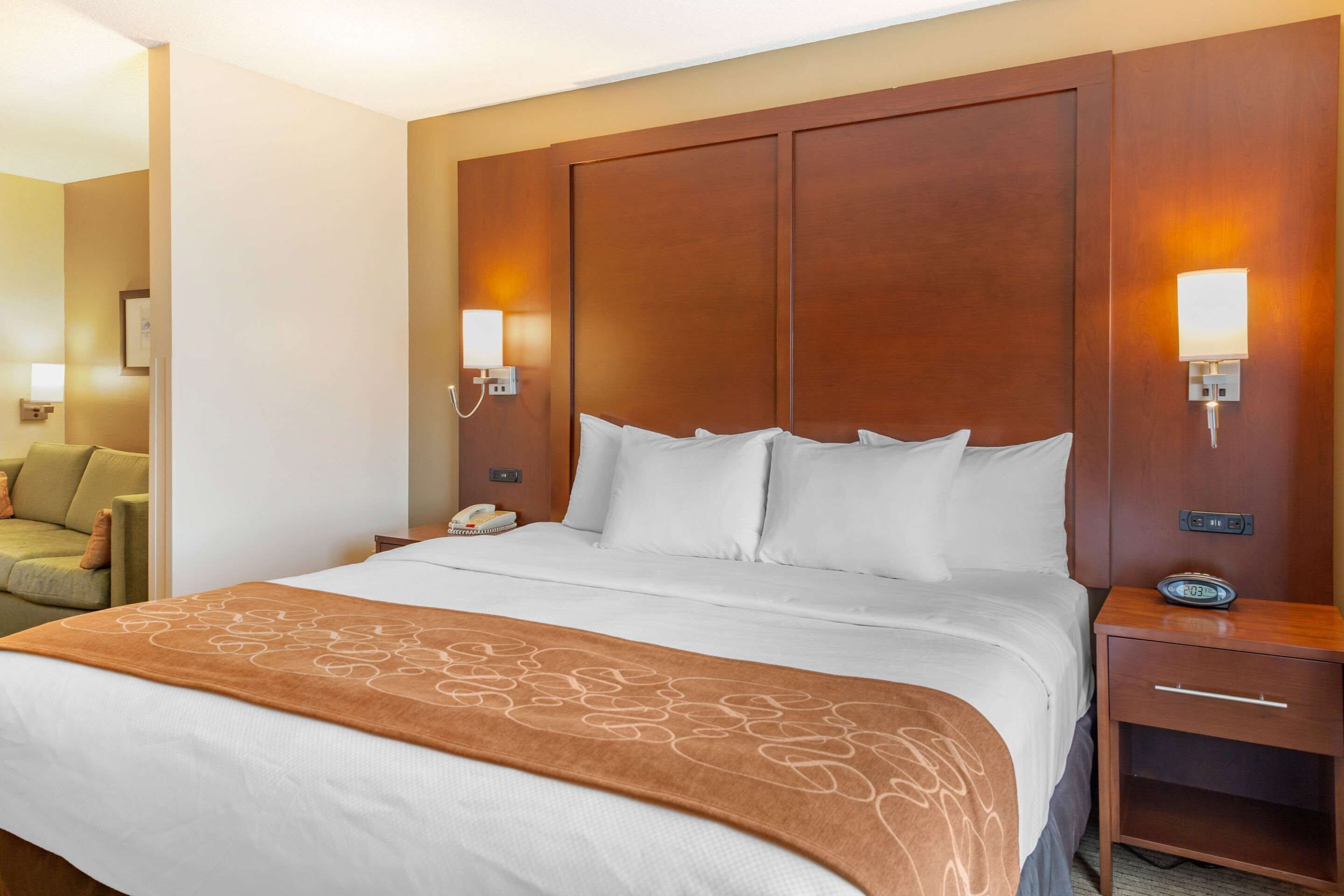 A hotel room with a large bed, a wooden headboard, bedside lamps, a nightstand, and a chair in the corner.