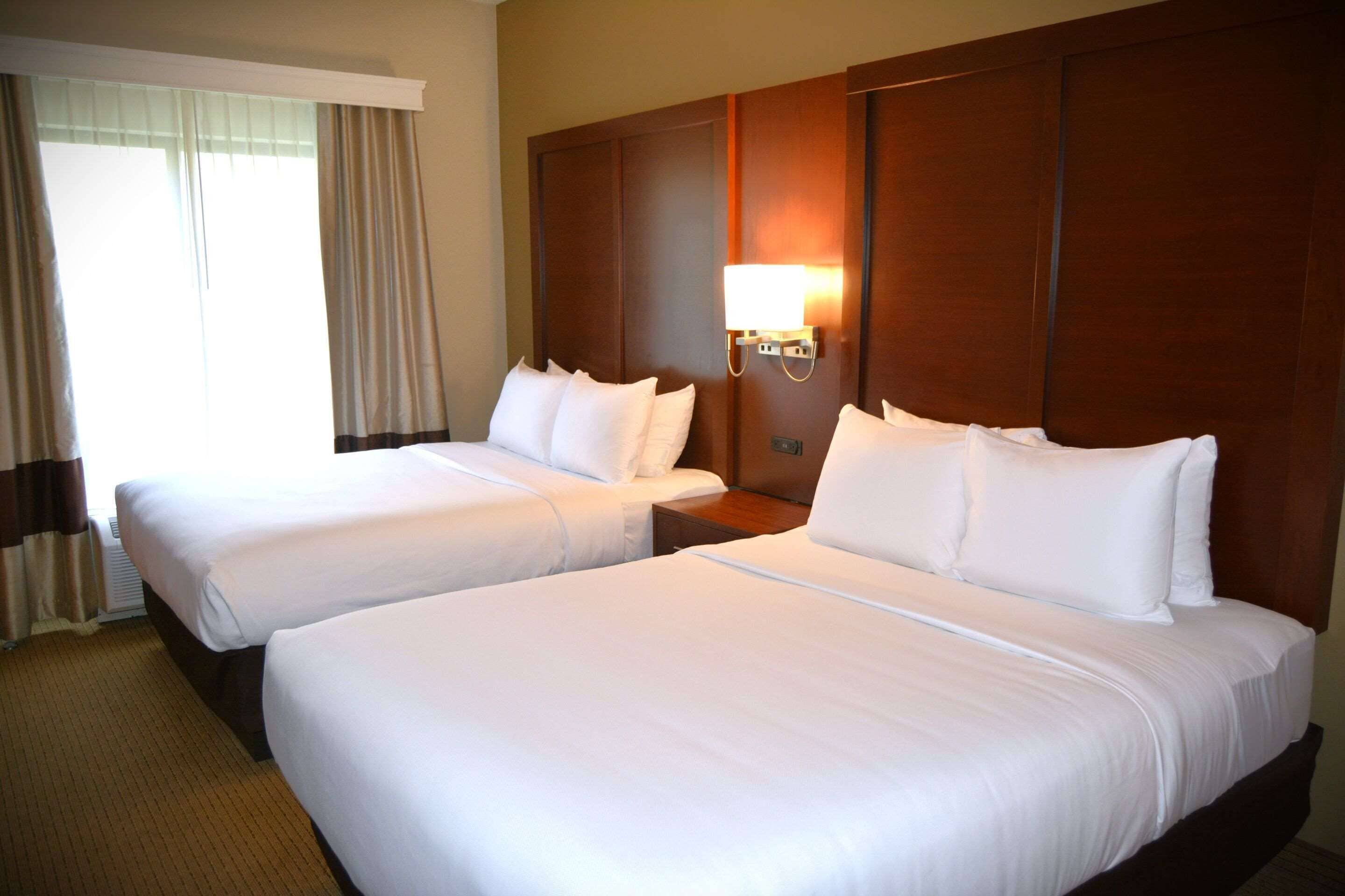 Habitación de hotel con dos camas individuales, una mesita de noche, una lámpara de pared y un ventanal con cortinas.