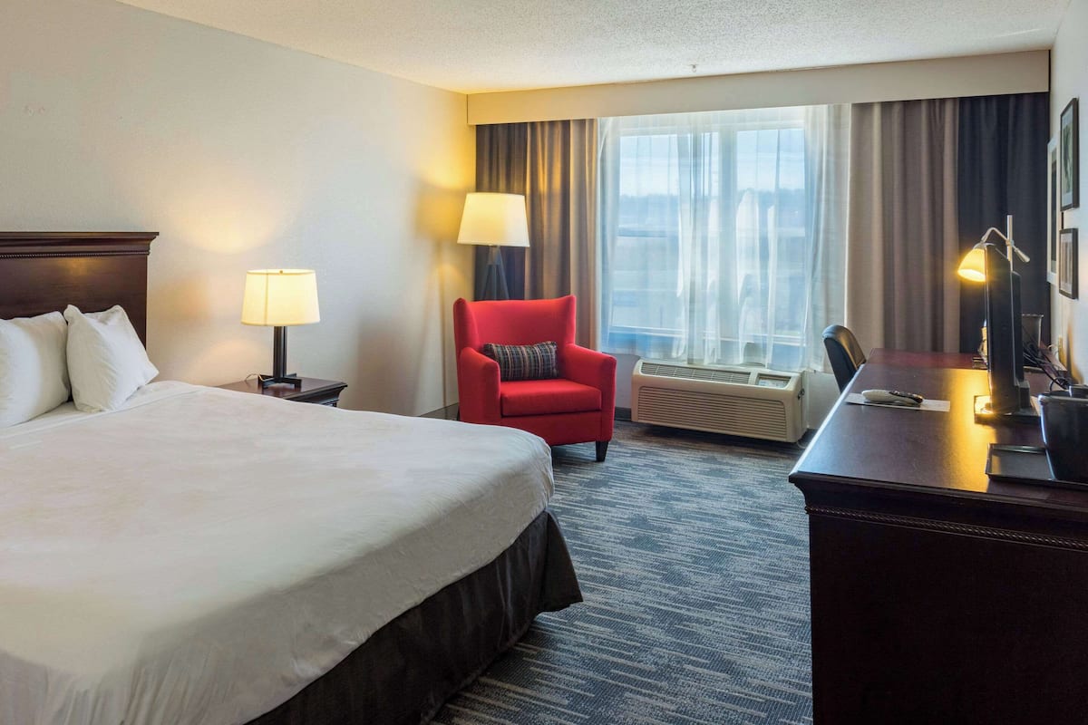A hotel room with a large bed, a desk, a red armchair, and a view of the city through the window.
