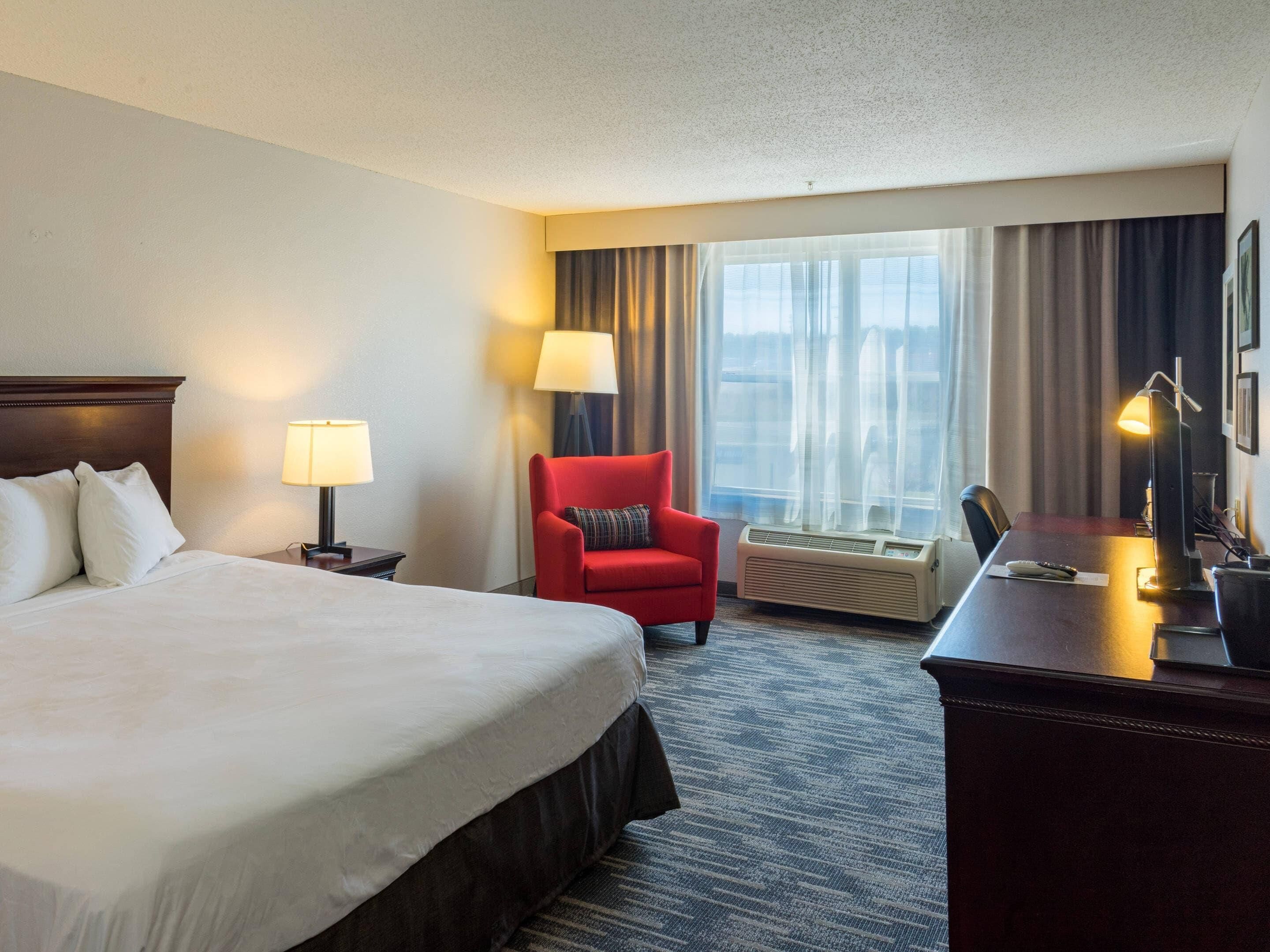 A hotel room with a large bed, a desk with a lamp, a red armchair, and a large window with curtains.