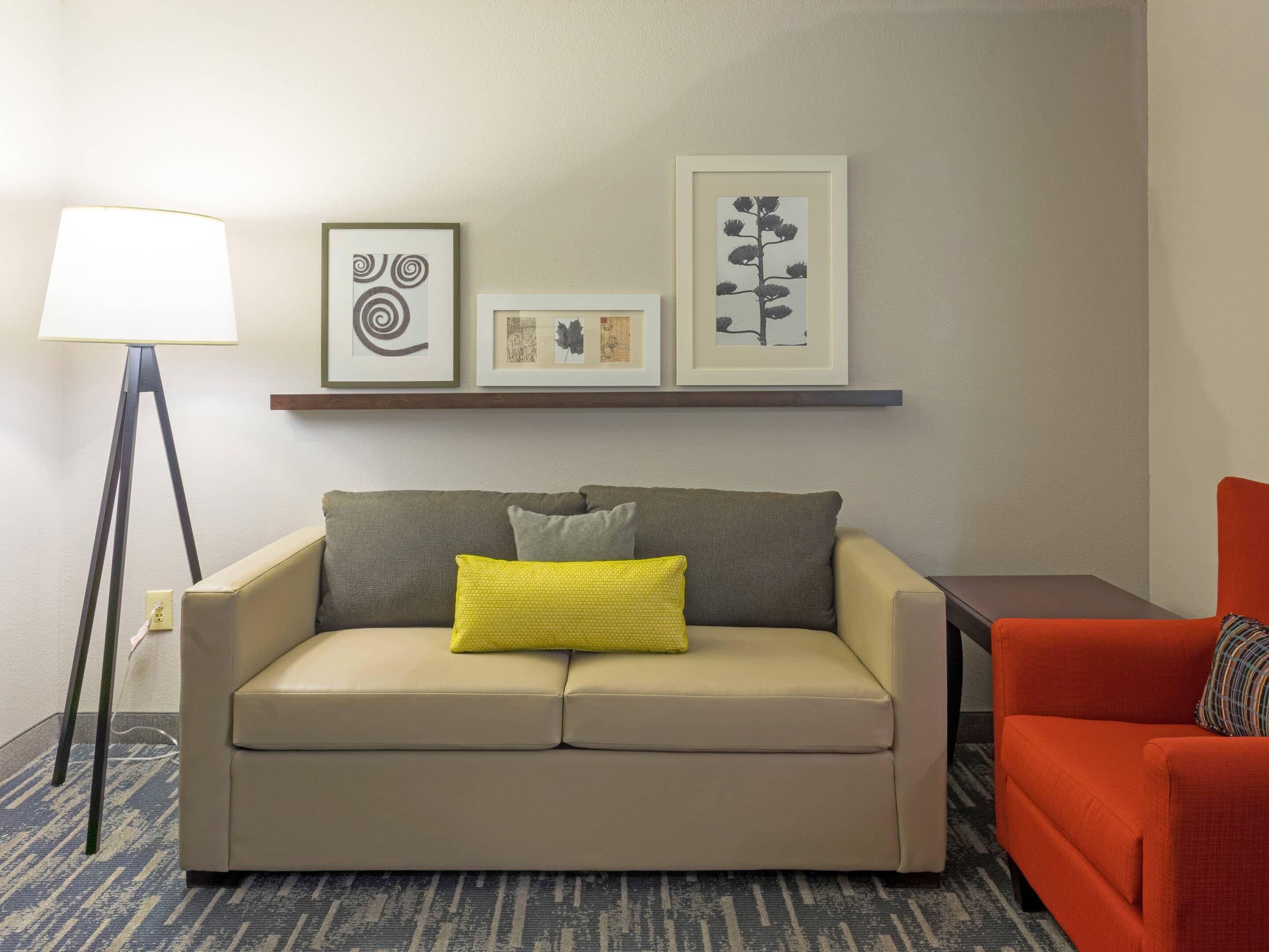 A neutral-toned living room with a beige sofa, a yellow pillow, an orange armchair, and a floor lamp. There are framed artworks on the wall above the sofa.