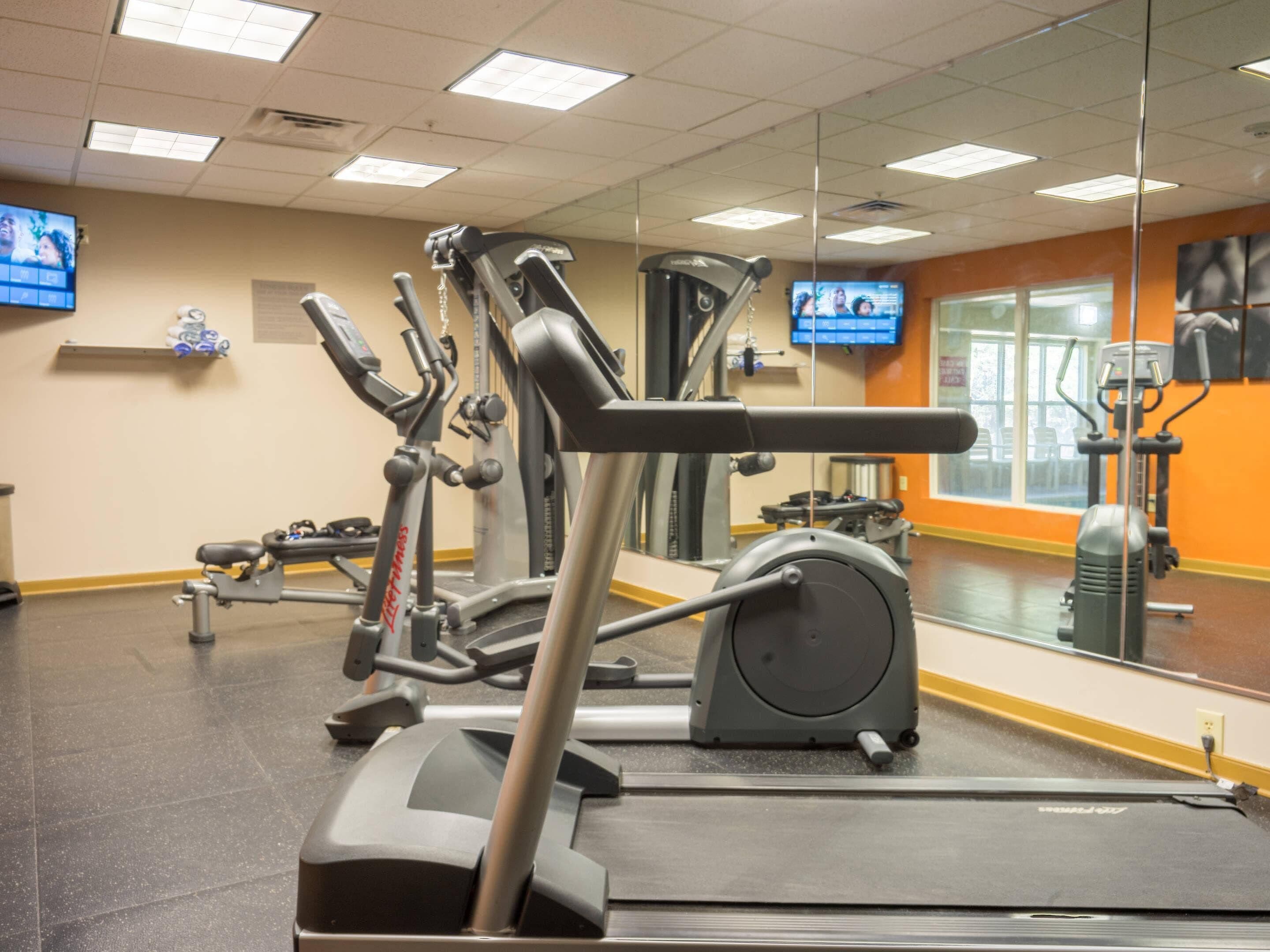 Un gimnasio con equipo de ejercicio, un muro espejado y un televisor en la pared.