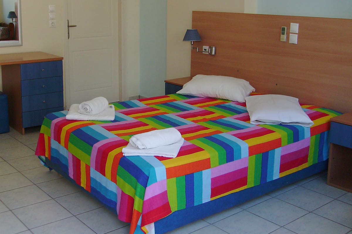 A bed with a colorful striped bedspread and white pillows, a wooden headboard, a bedside table with a lamp, and a blue dresser.