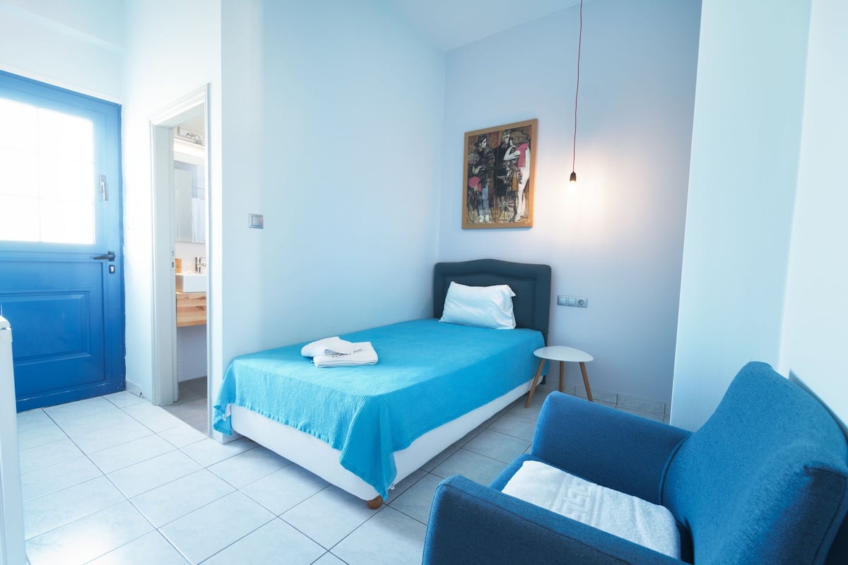 A bedroom with a bed, a blue chair, a bedside table, and a painting on the wall.