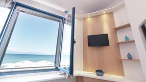 A room with a large window offering a view of the sea, a mounted TV on the wall, and wooden shelves with decorative items. - Aggelos Hotel (Messini)