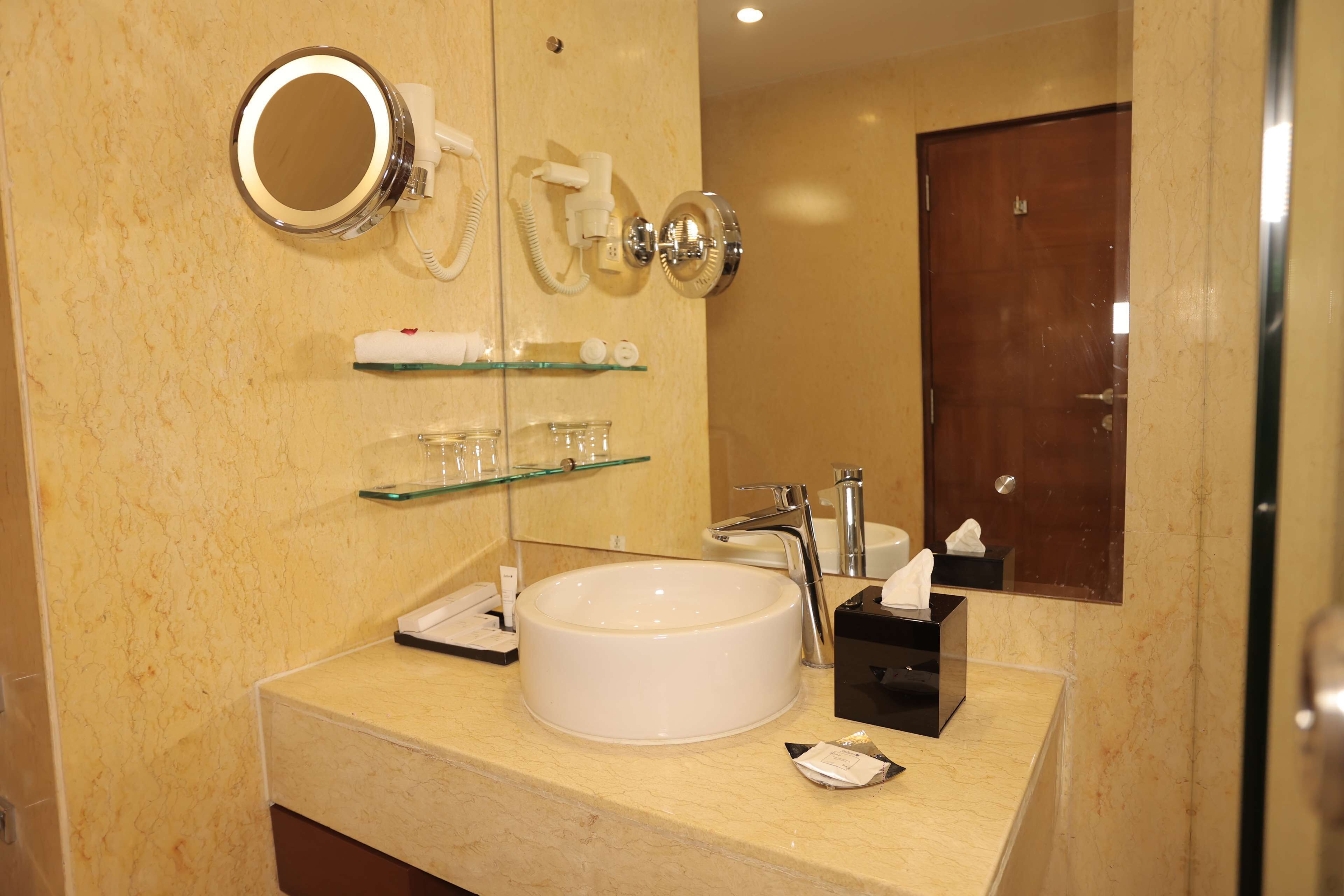 A bathroom with a round sink, a mirror, a glass shelf with toiletries, and a door in the background.