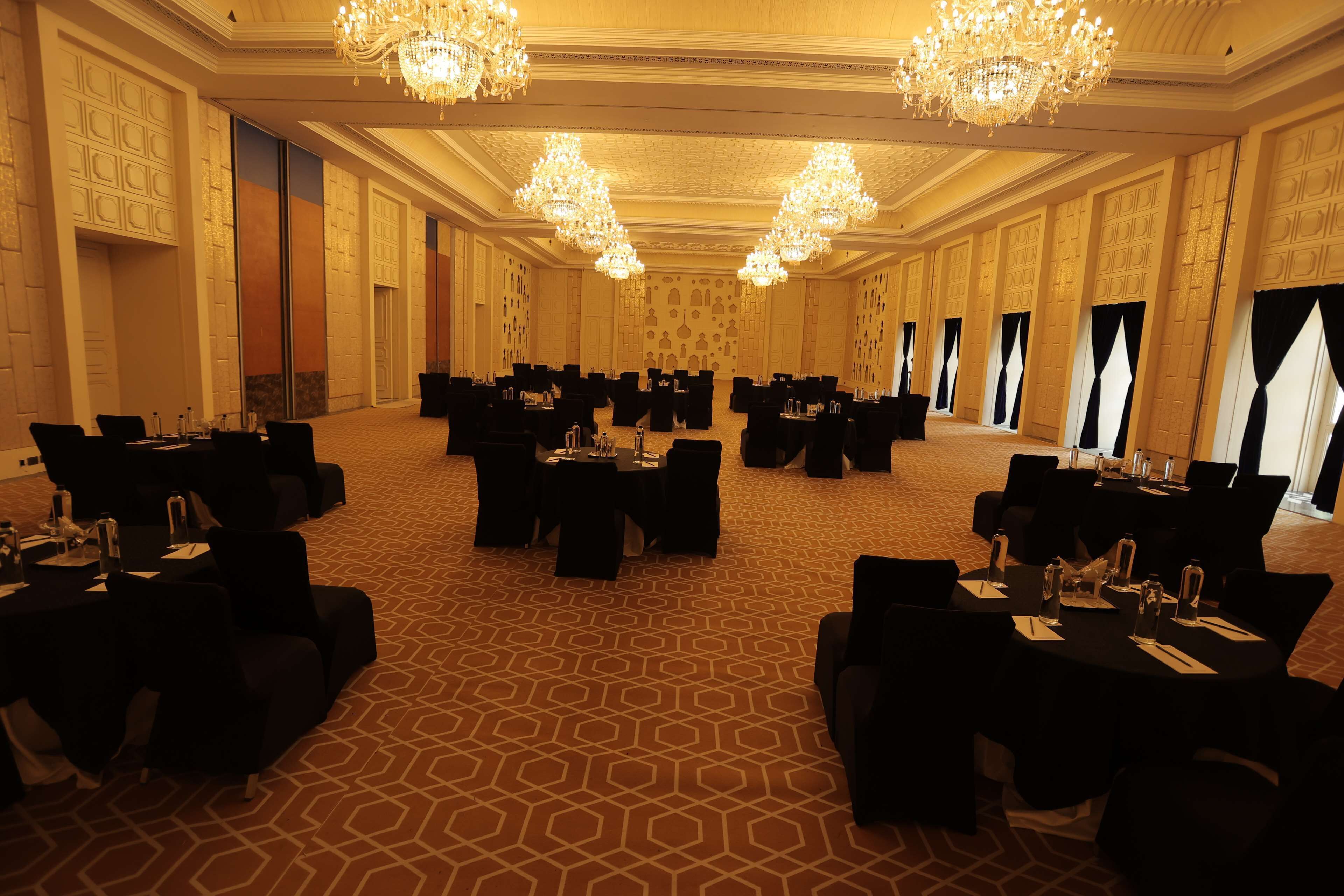 A spacious banquet hall with multiple round tables and chairs, ornate chandeliers, and patterned carpet.