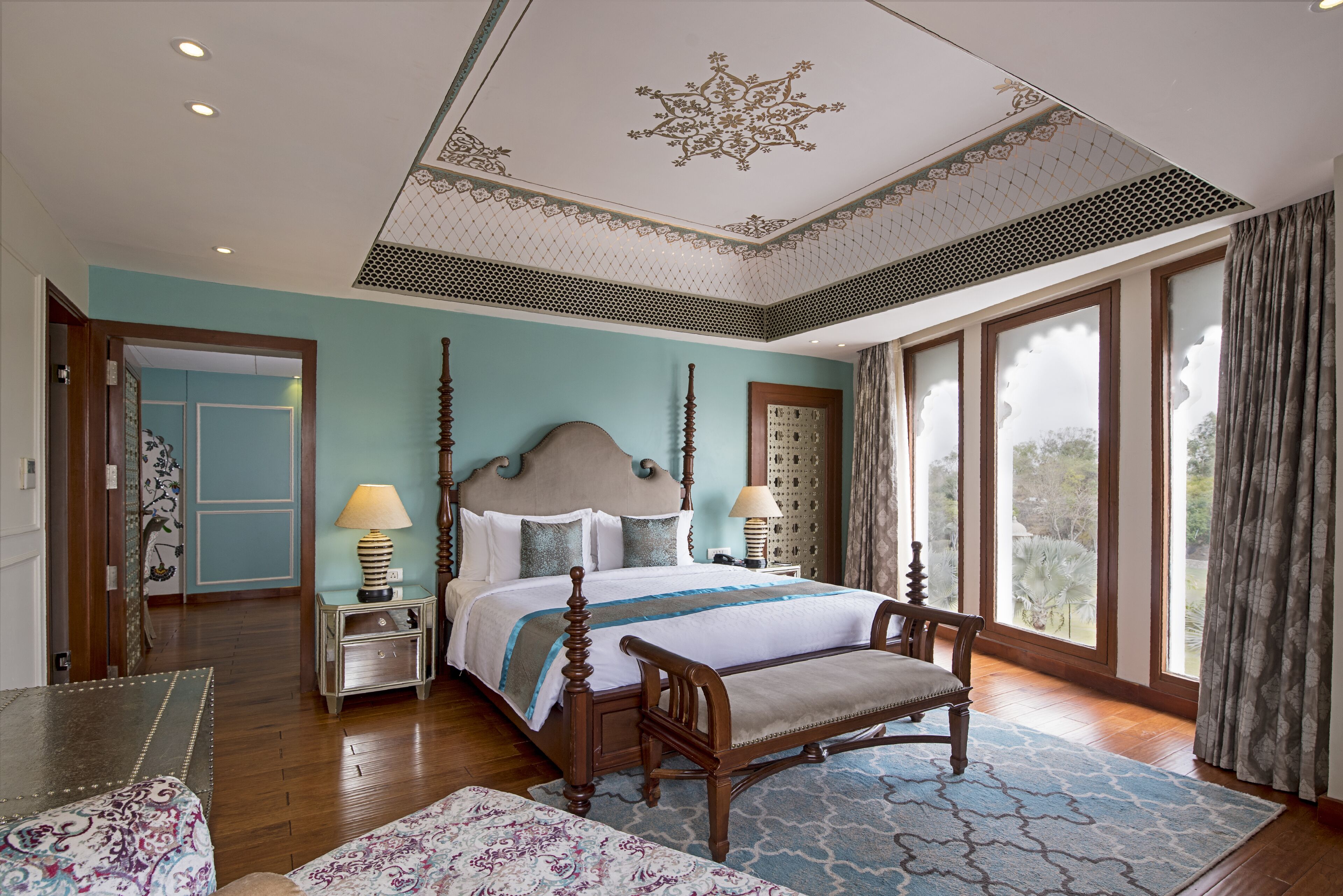A spacious bedroom with a large bed, a wooden bench, and a view of the outdoors through large windows.