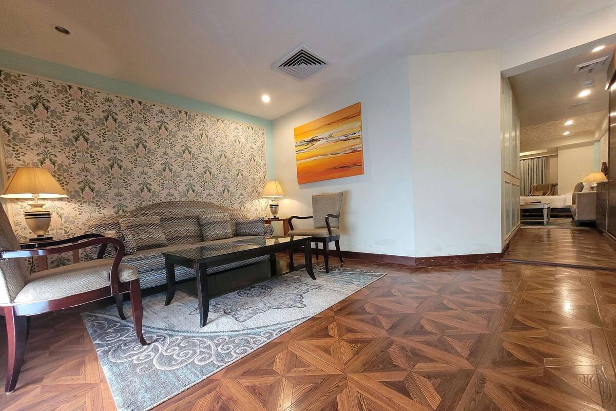 Ein Wohnzimmer mit ornamentalem Wandpapier, einer Couch, zwei Sesseln, einem Couchtisch und einem Teppich.