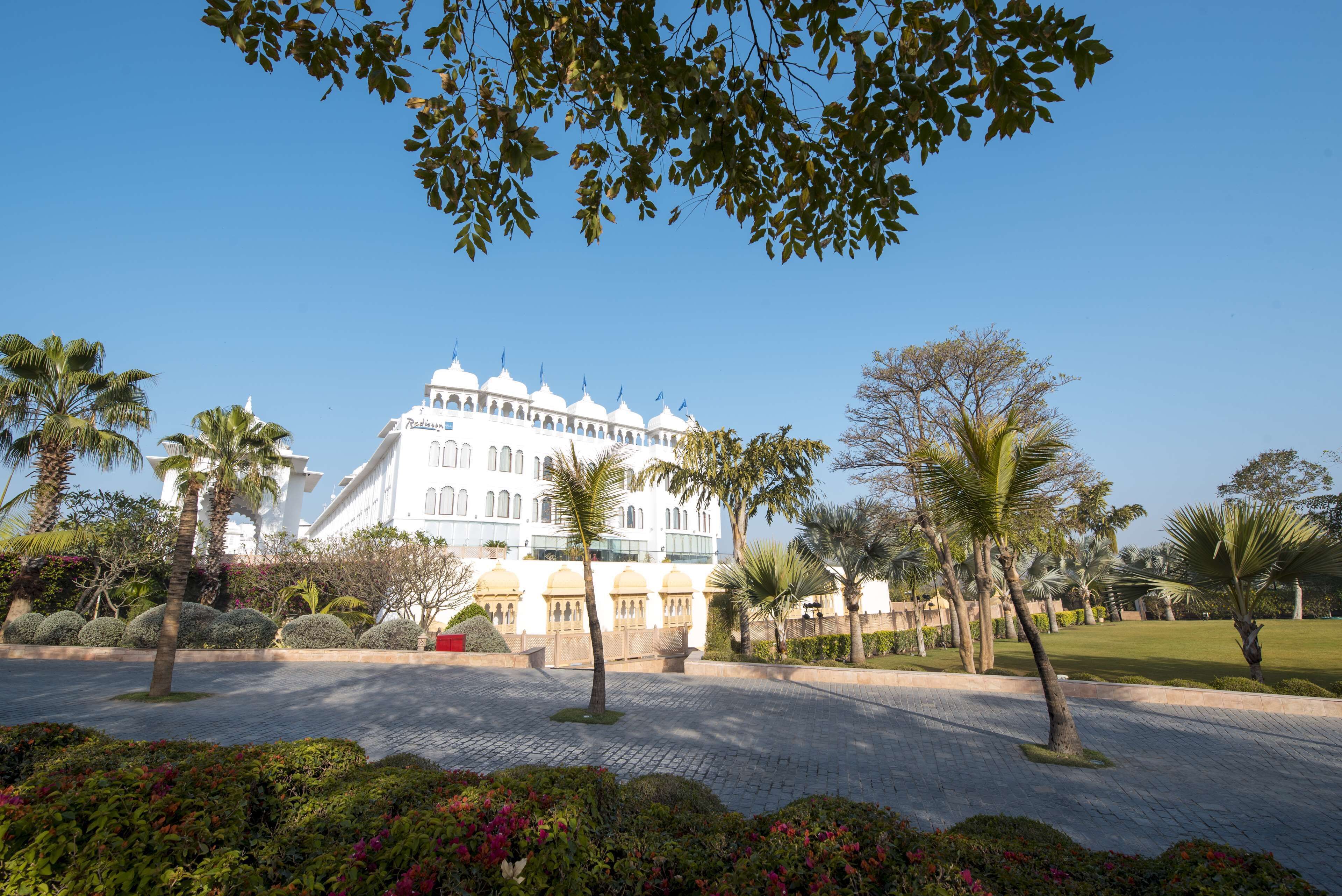 Un grand bâtiment blanc, doté de nombreuses fenêtres et surmonté d’un dôme central, entouré de palmiers et de jardins bien entretenus.