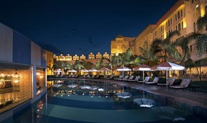 A poolside area with lounge chairs and umbrellas, a large building with domed structures in the background, and a clear night sky.