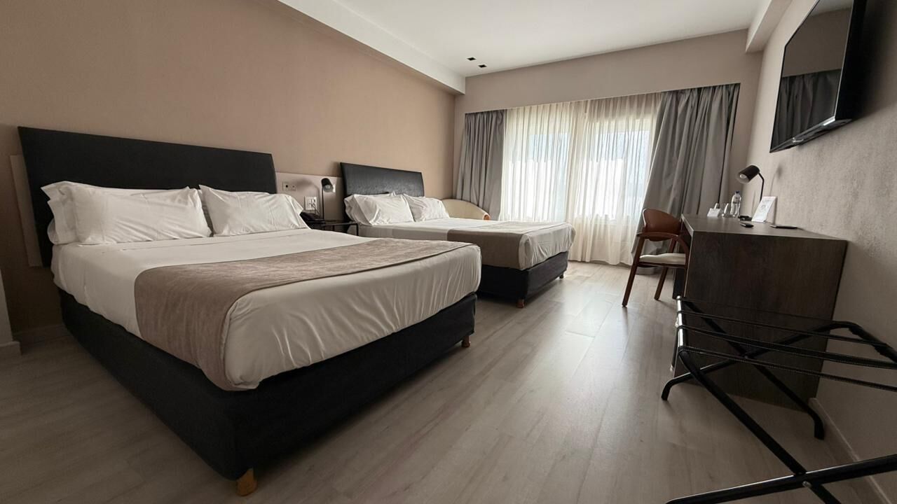 Habitación de hotel con dos camas, un escritorio, una silla, un televisor y un ventanal grande con cortinas.