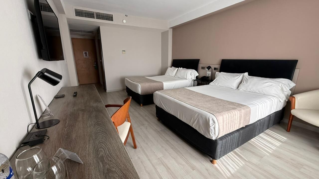 A hotel room with two beds, a desk, a chair, a TV, and a door.