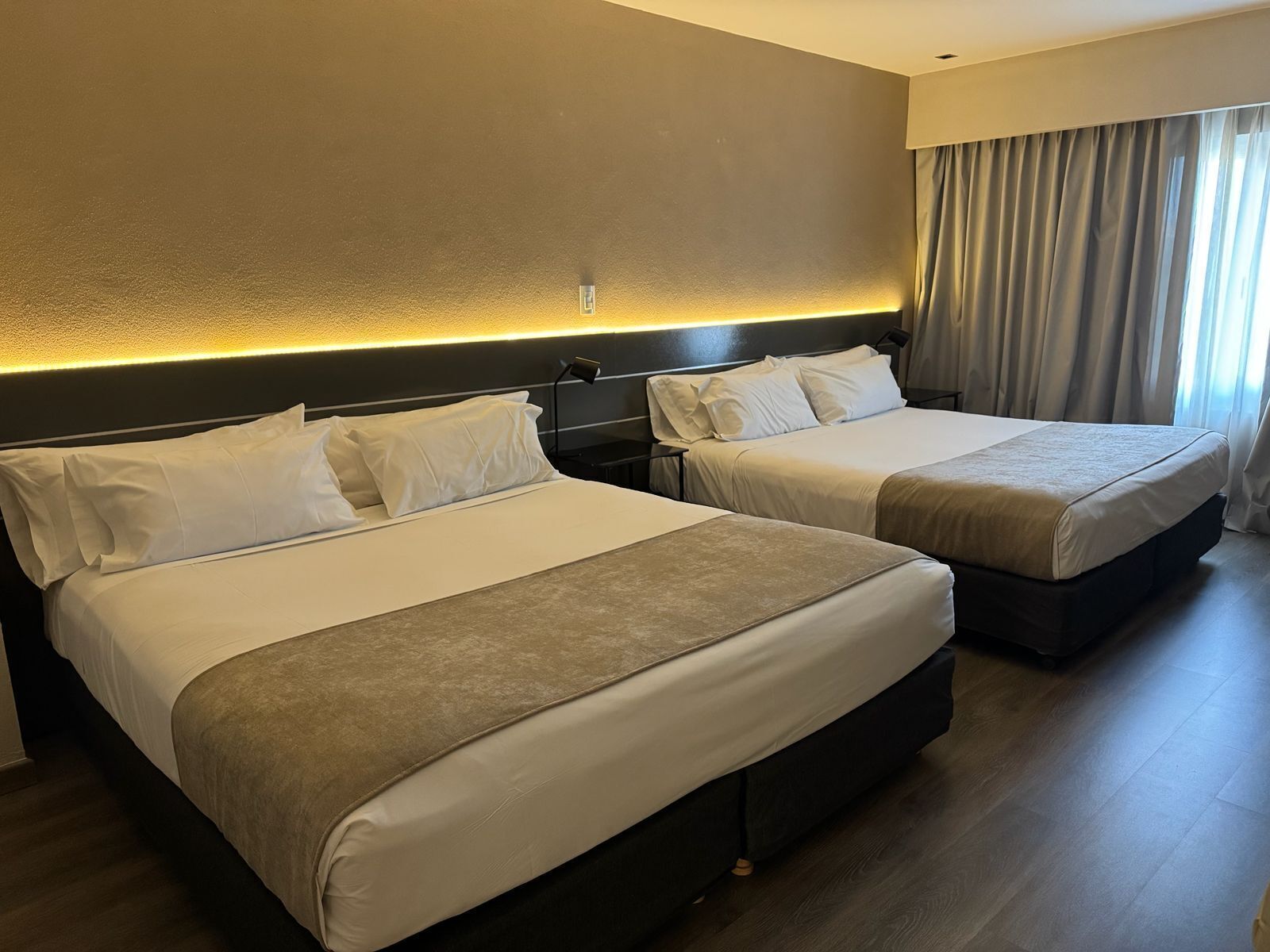 A hotel room with two beds, a headboard with built-in lighting, a nightstand, and a window with curtains.