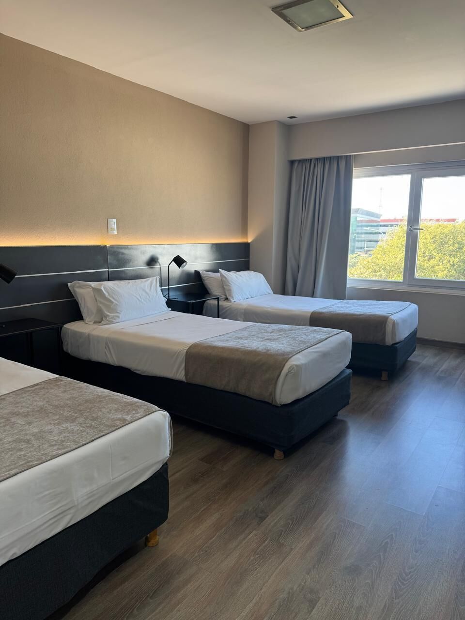 Habitación de hotel con dos camas, un ventanal con cortinas y vista a espacios verdes.