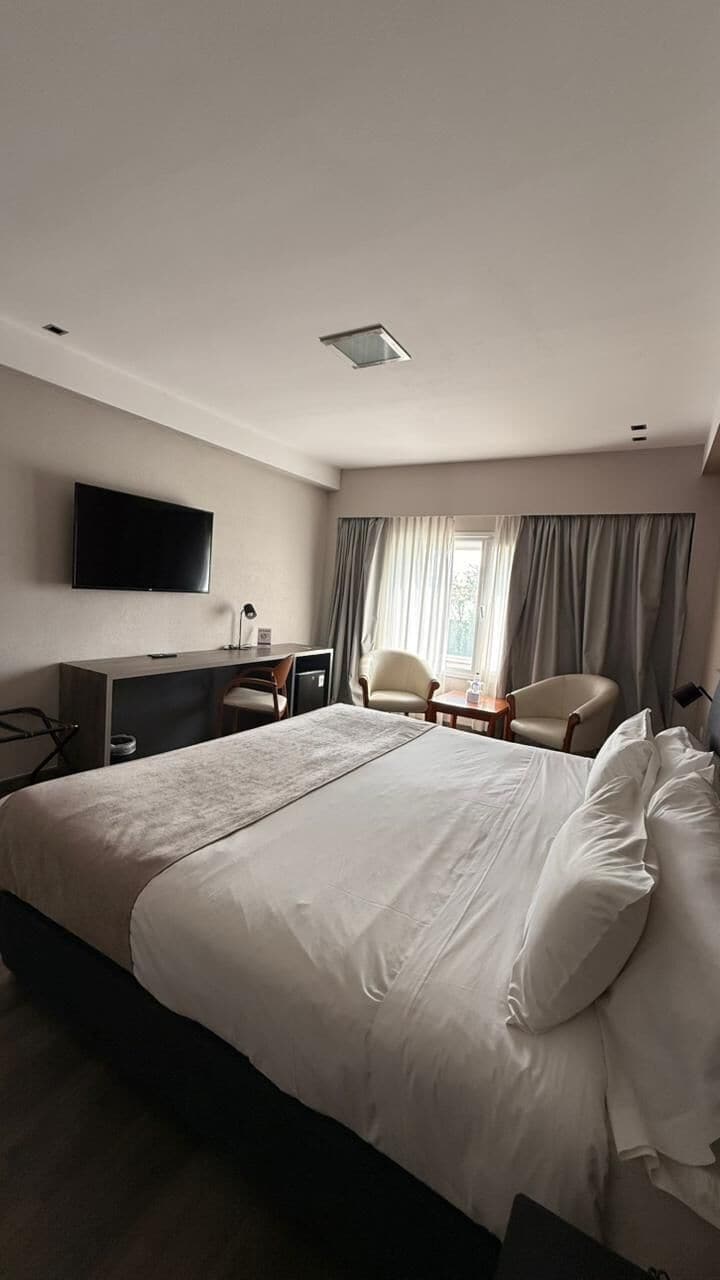 A hotel room with a large bed, a desk, a chair, a TV, and a window with curtains.