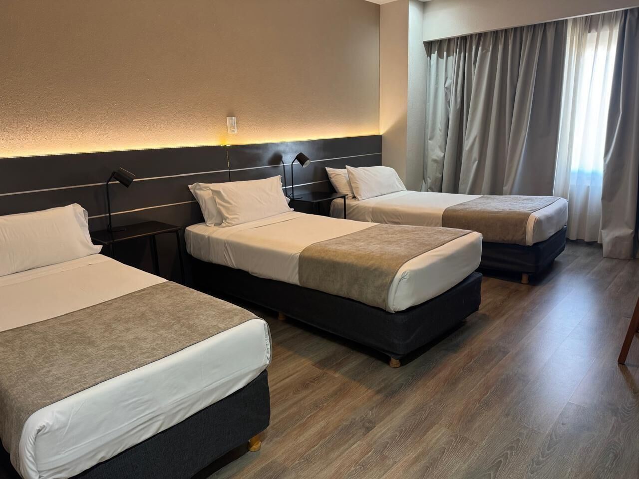 Habitación de hotel con tres camas, una mesita de noche con lámpara y un ventanal con cortinas.