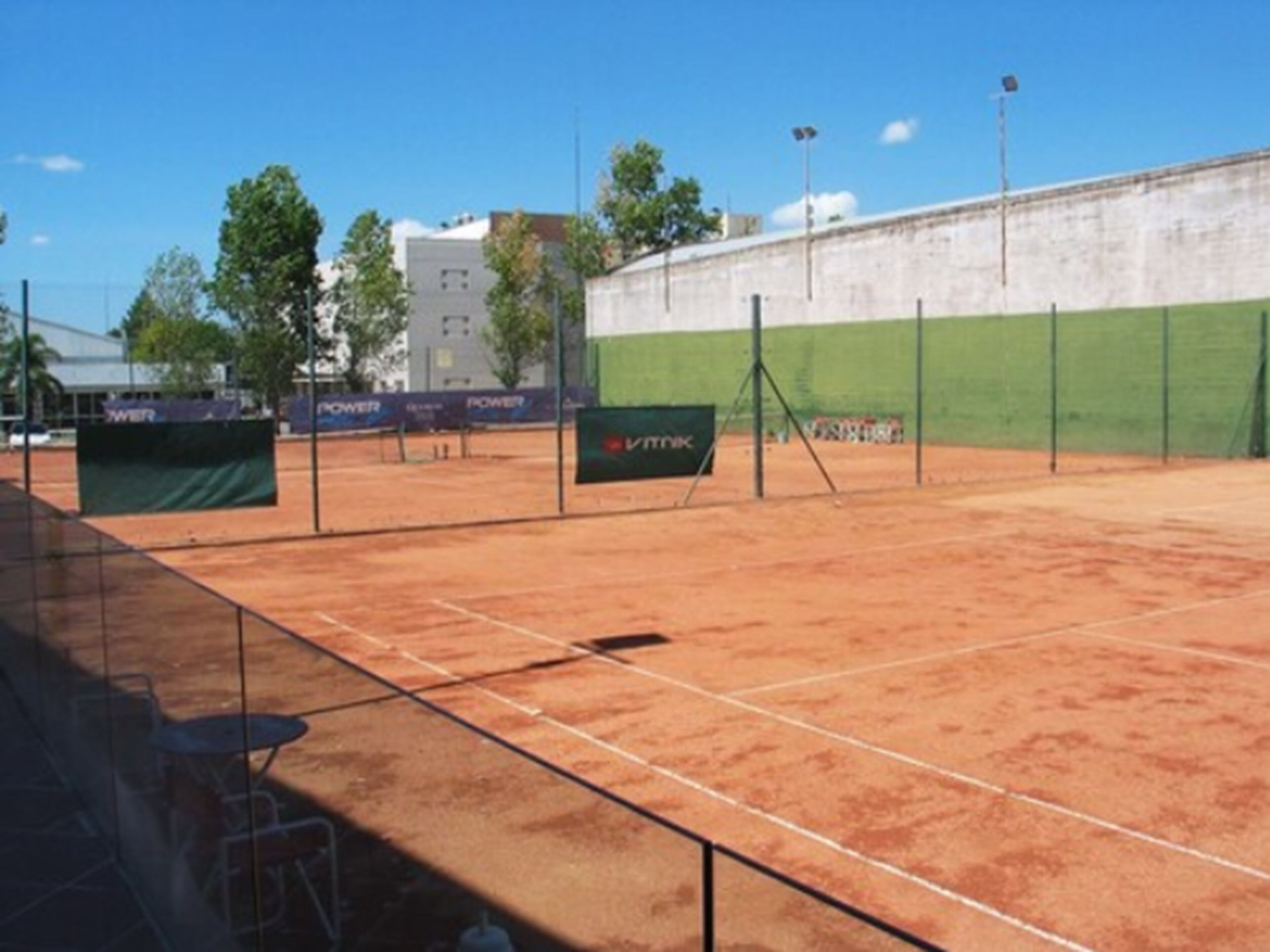 An outdoor tennis court with a green fence, a dirt surface, and a white wall in the background.