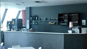 Un restaurante moderno con mesas y sillas blancas, una zona de bar con botellas y copas de vino, y un amplio ventanal que proporciona luz natural.