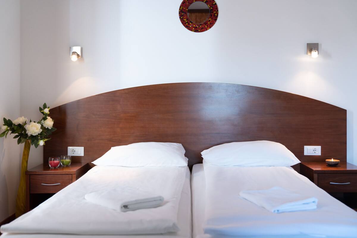 Habitación de hotel con dos camas, cabecero de madera, mesitas de noche con lámparas, un jarrón con flores y un espejo en la pared.