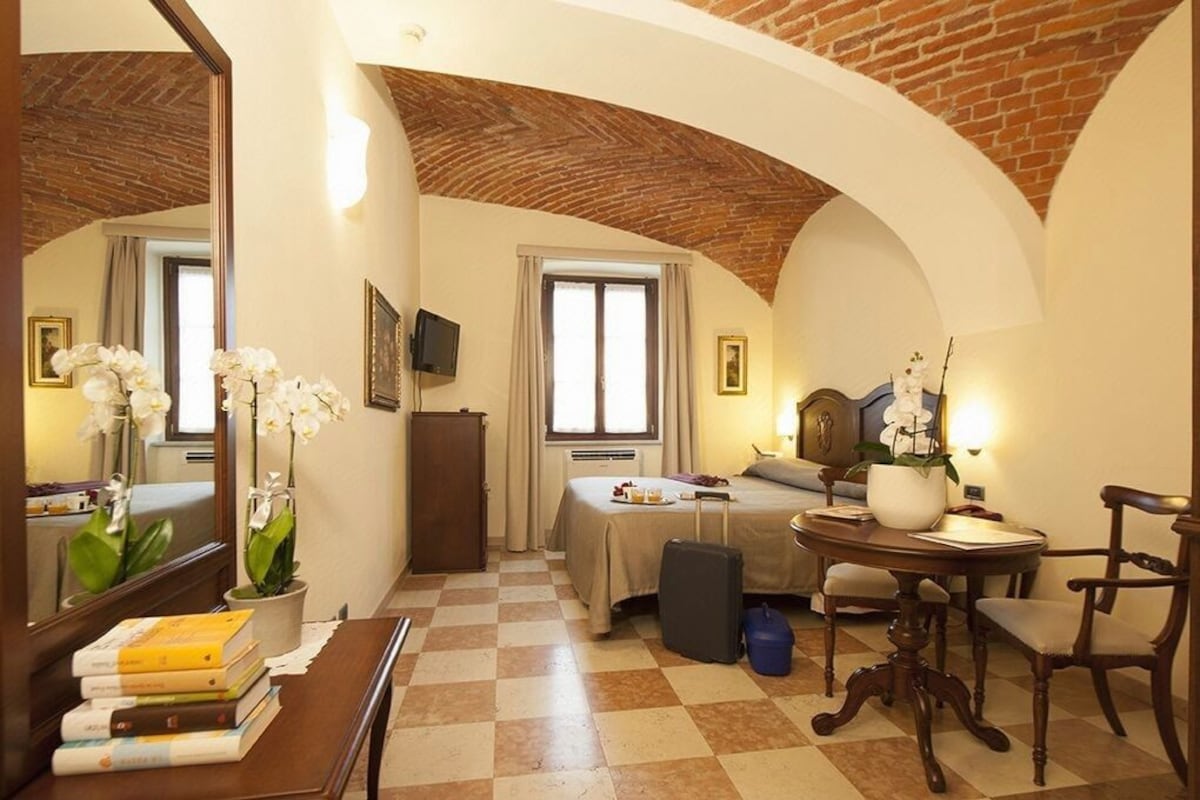 A hotel room with a checkered floor, a bed, a desk with books, a mirror, and a potted plant.