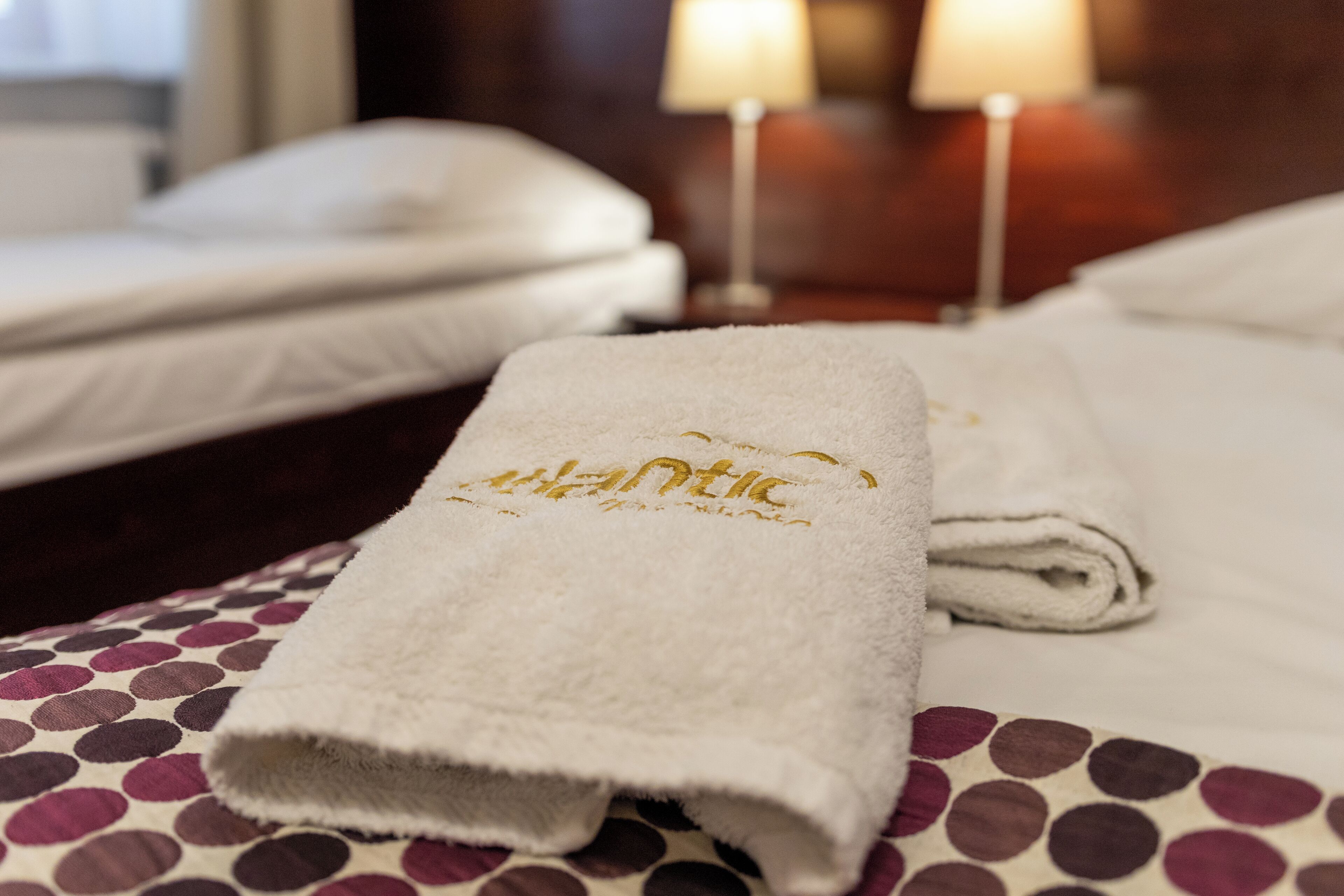 Una cama bien hecha con una toalla blanca que tiene letras doradas, una colcha estampada de lunares y una mesita de noche de madera con dos lámparas.