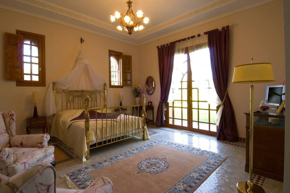 A bedroom with a four-poster bed, a chandelier, a desk with a lamp, and a view of the outdoors through a large window.