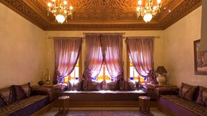 A traditional room with ornate wooden ceiling, a large ornate rug, and a seating area with cushions and a bench. - Dar Kantzaro (Marrakech)