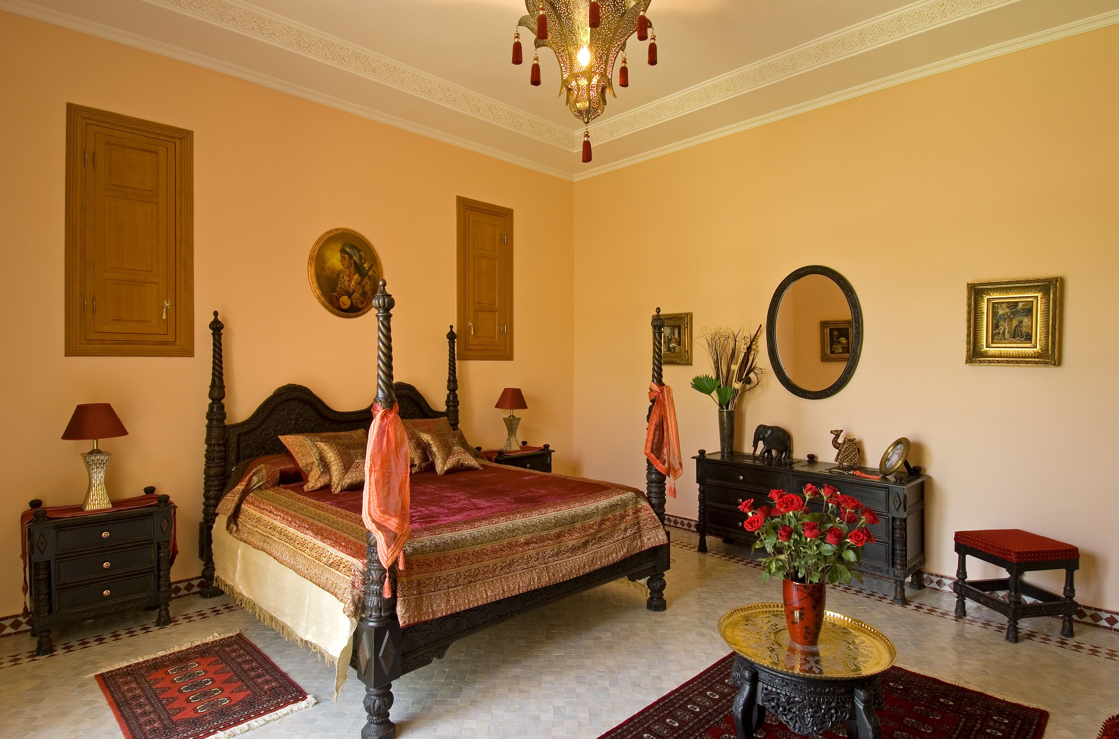 A traditional four-poster bed with a red and gold bedspread, a chandelier, a mirror, and a vase with red flowers on a round table.
