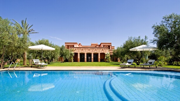 A swimming pool with tiled flooring, surrounded by lounge chairs and umbrellas, with a building in the background. - Dar Kantzaro (Marrakech)