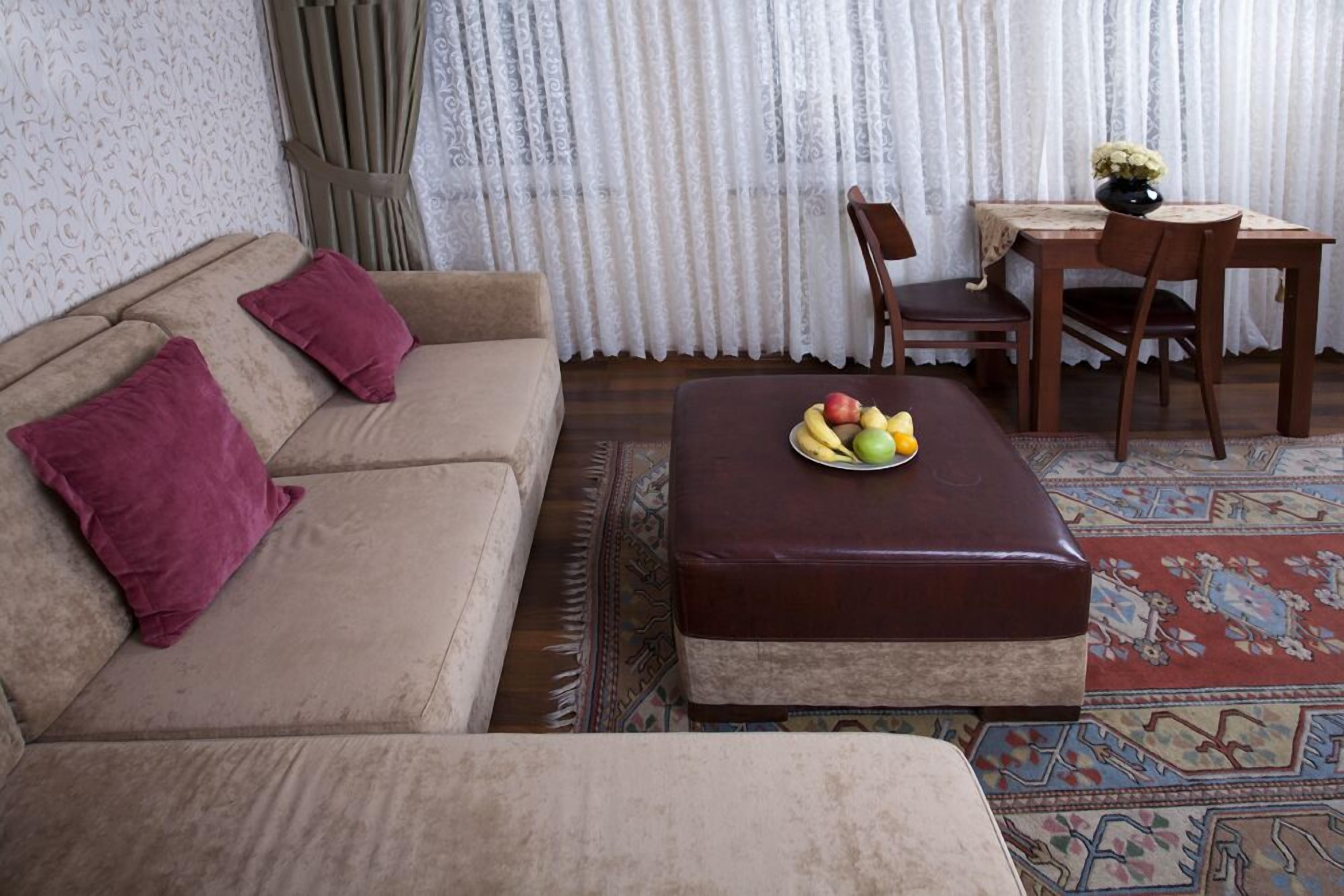 A living room with a beige sofa, two red cushions, a brown ottoman with a fruit bowl, a wooden dining table with chairs, and a patterned rug.