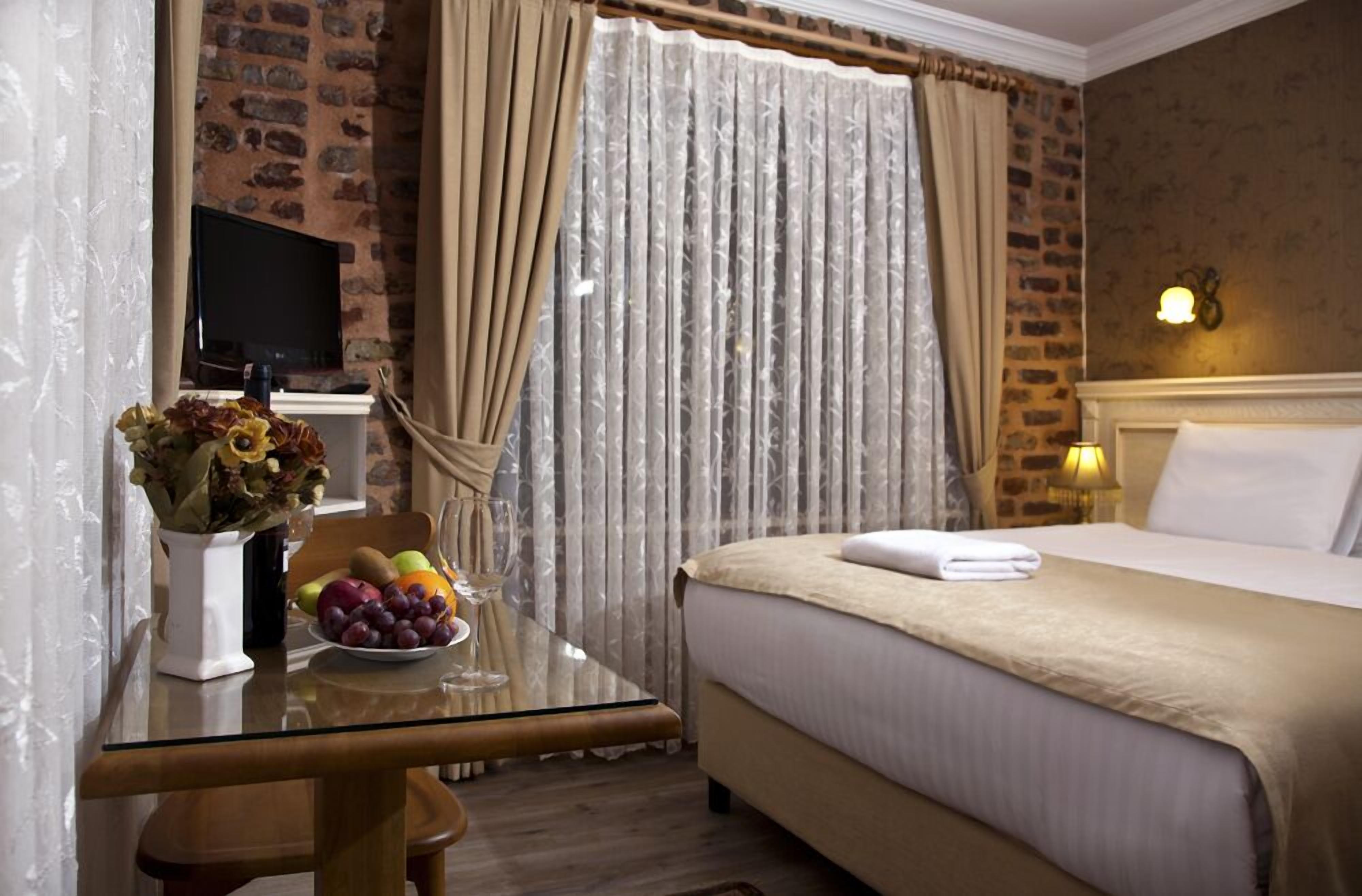 Una habitación de hotel con cama, una mesa de vidrio con fruta y un jarrón, cortinas y una pared de ladrillo.