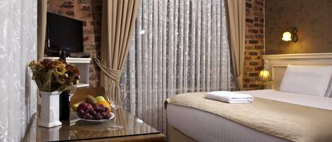 Una habitación de hotel con cama, una mesa de vidrio con fruta y un jarrón, cortinas y una pared de ladrillo.